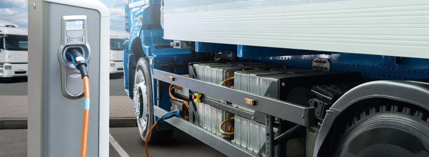 An electric truck charging at a depot, with its battery pack visible beneath the chassis.