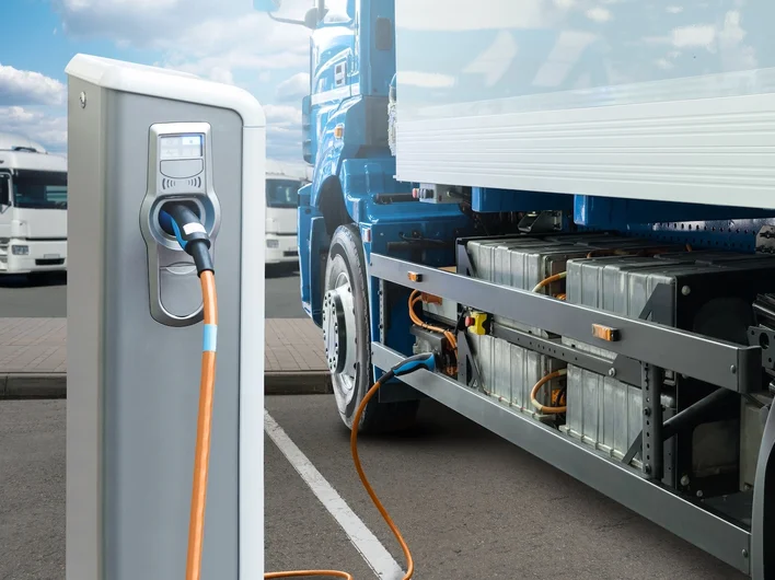 An electric truck charging at a depot, with its battery pack visible beneath the chassis.